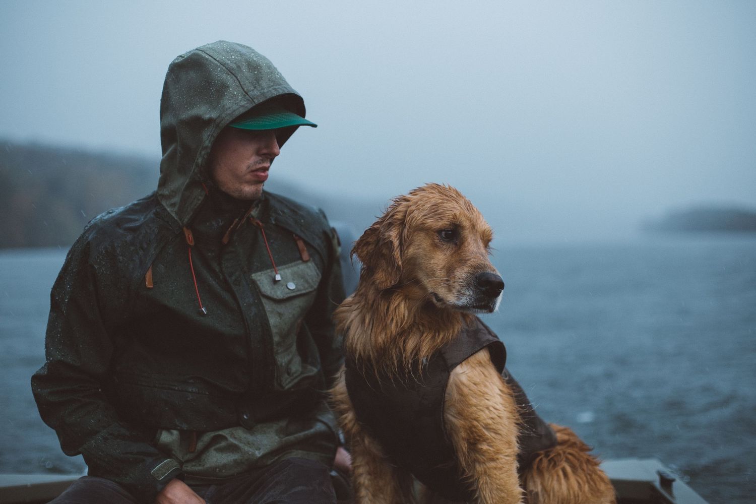 Man in rain jacket sits with wet golden retriever dog on boat in foggy misty lake landscape.