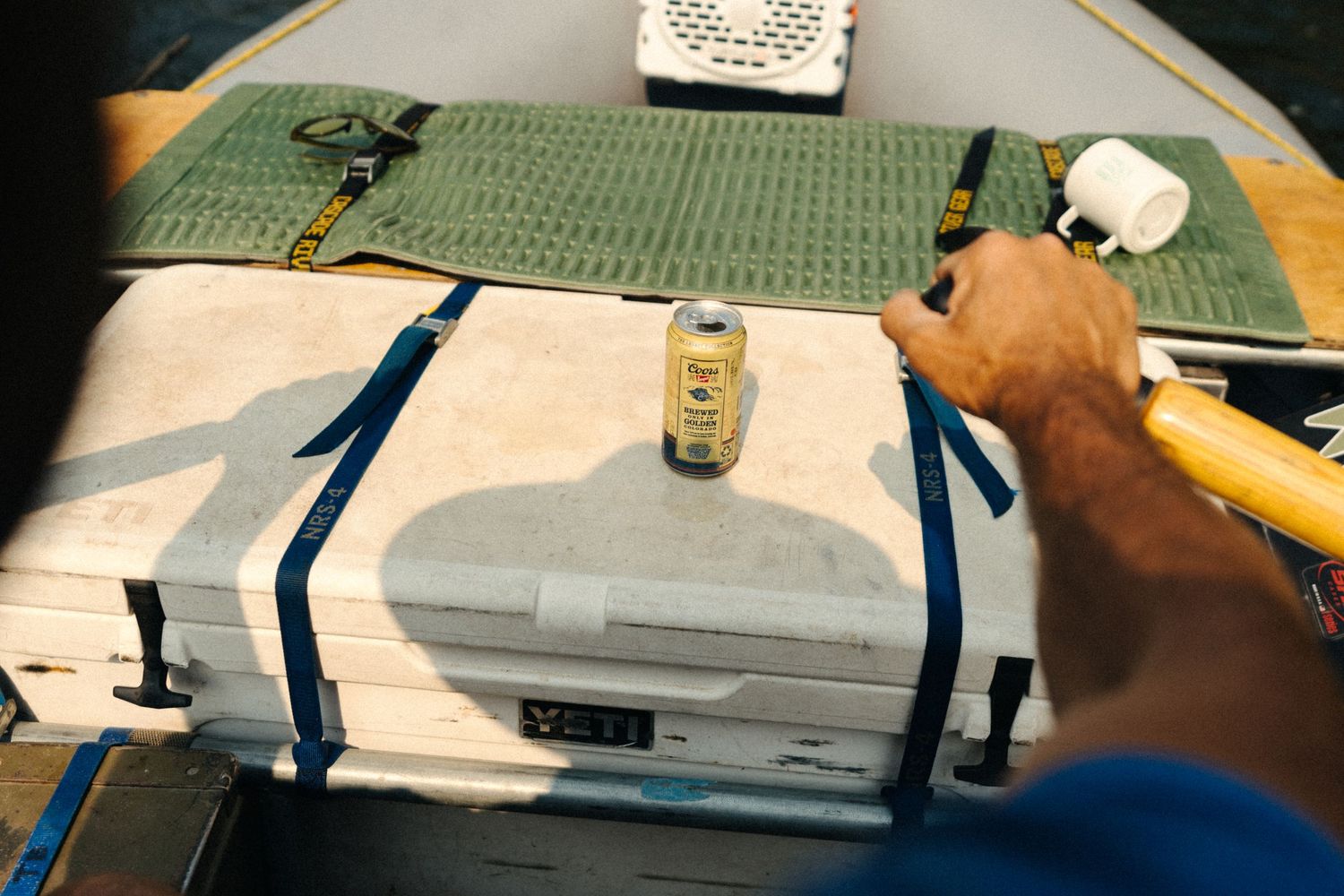Man securing a YETI cooler with blue straps on a boat deck, with a can on top and fishing gear nearby.