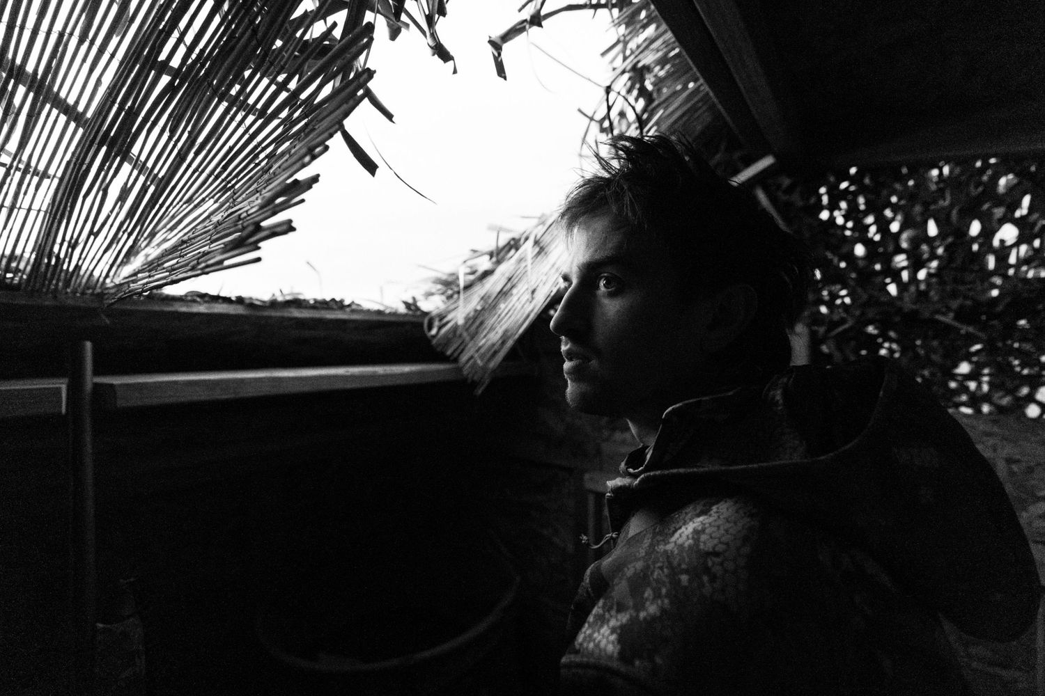 Black and white portrait of a pensive man sitting in shadow near a rustic thatched structure outdoors.