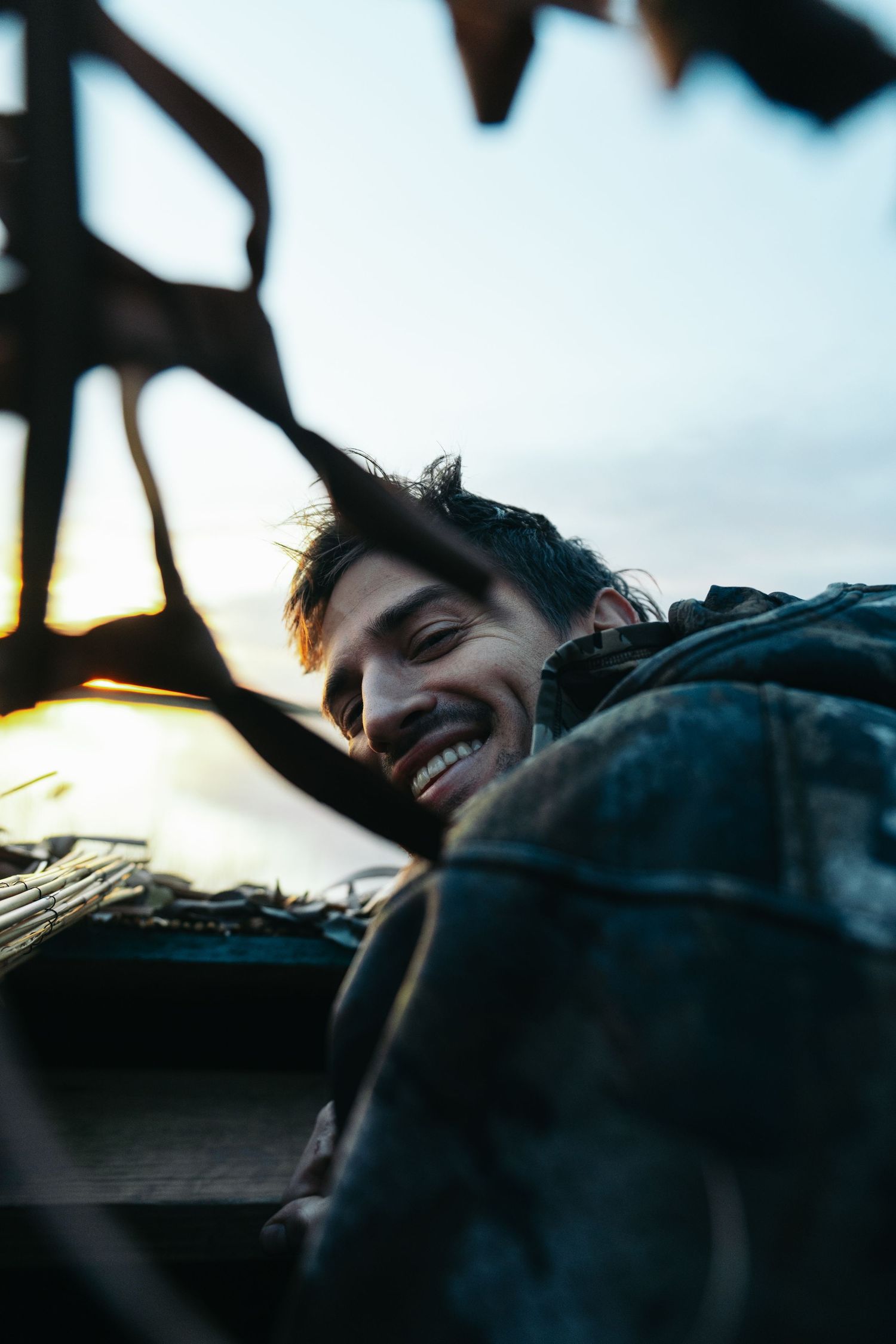 A smiling young man peers over a ledge at dusk, framed by dark silhouetted branches against a twilight sky.