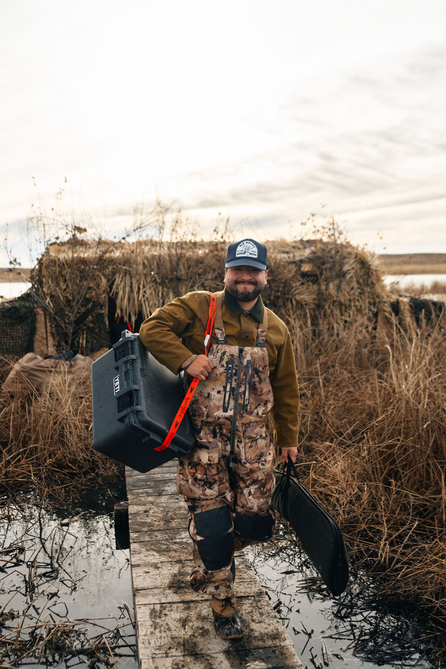Bearded hunter in camo waders carries a YETI cooler on a wooden dock surrounded by marshland.