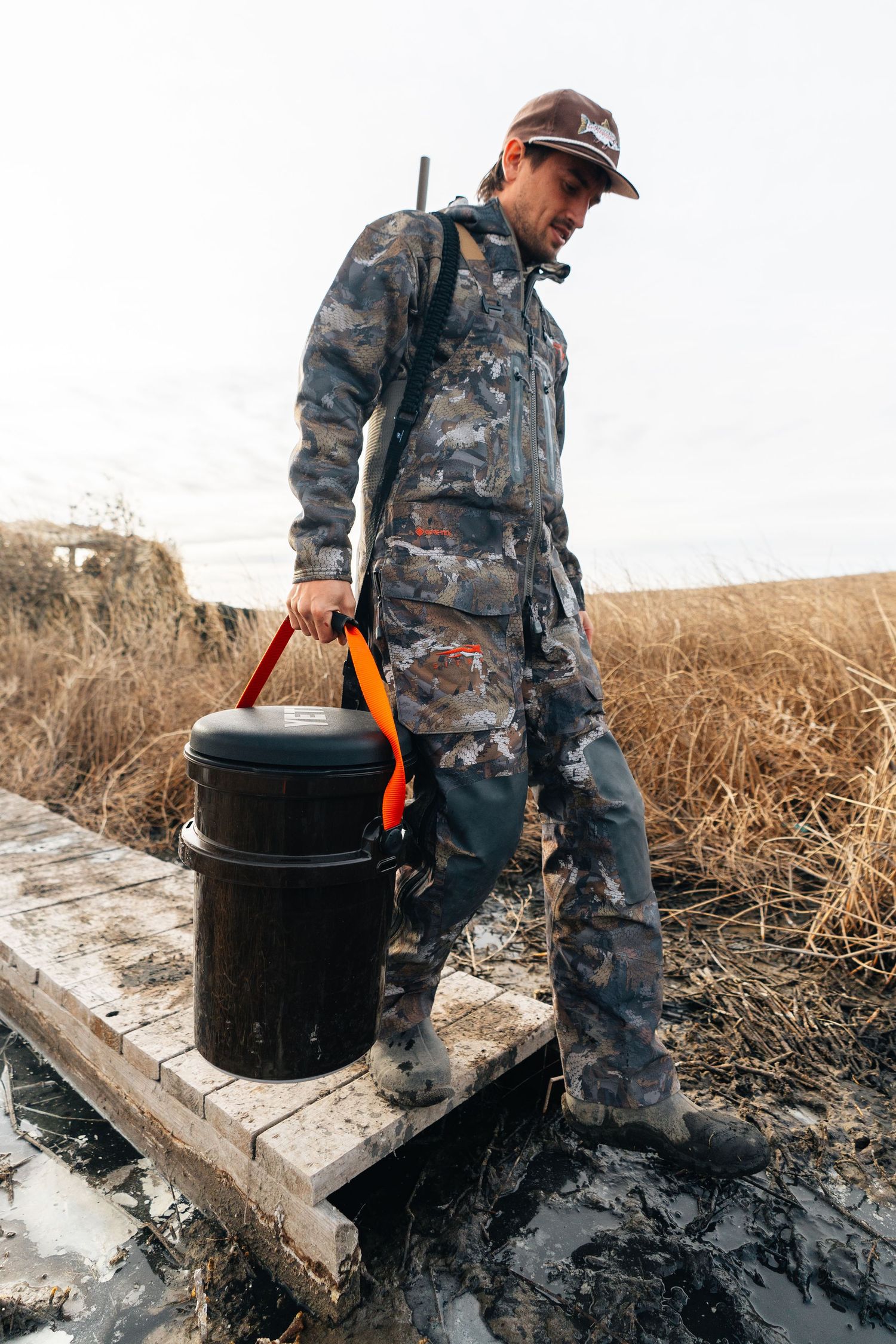 Hunter in camo gear carrying a black bucket on a wooden dock surrounded by marshland and dry grass.