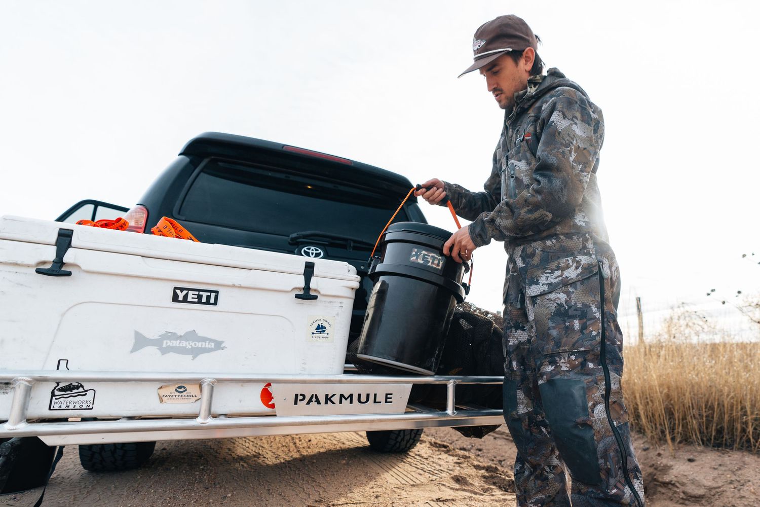 Hunter in camo gear loading a YETI cooler into a white PAKMULE truck bed outdoors.