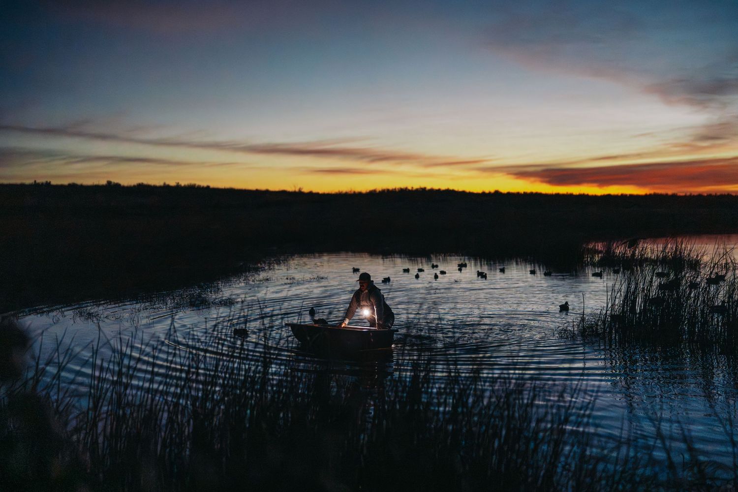Person in a small boat on a calm marsh at dusk, with a glowing light and silhouetted reeds against a colorful sunset sky.