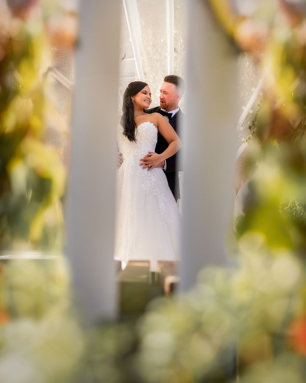 Bride in white gown and groom in tuxedo share romantic moment framed by white columns and lush floral decor.