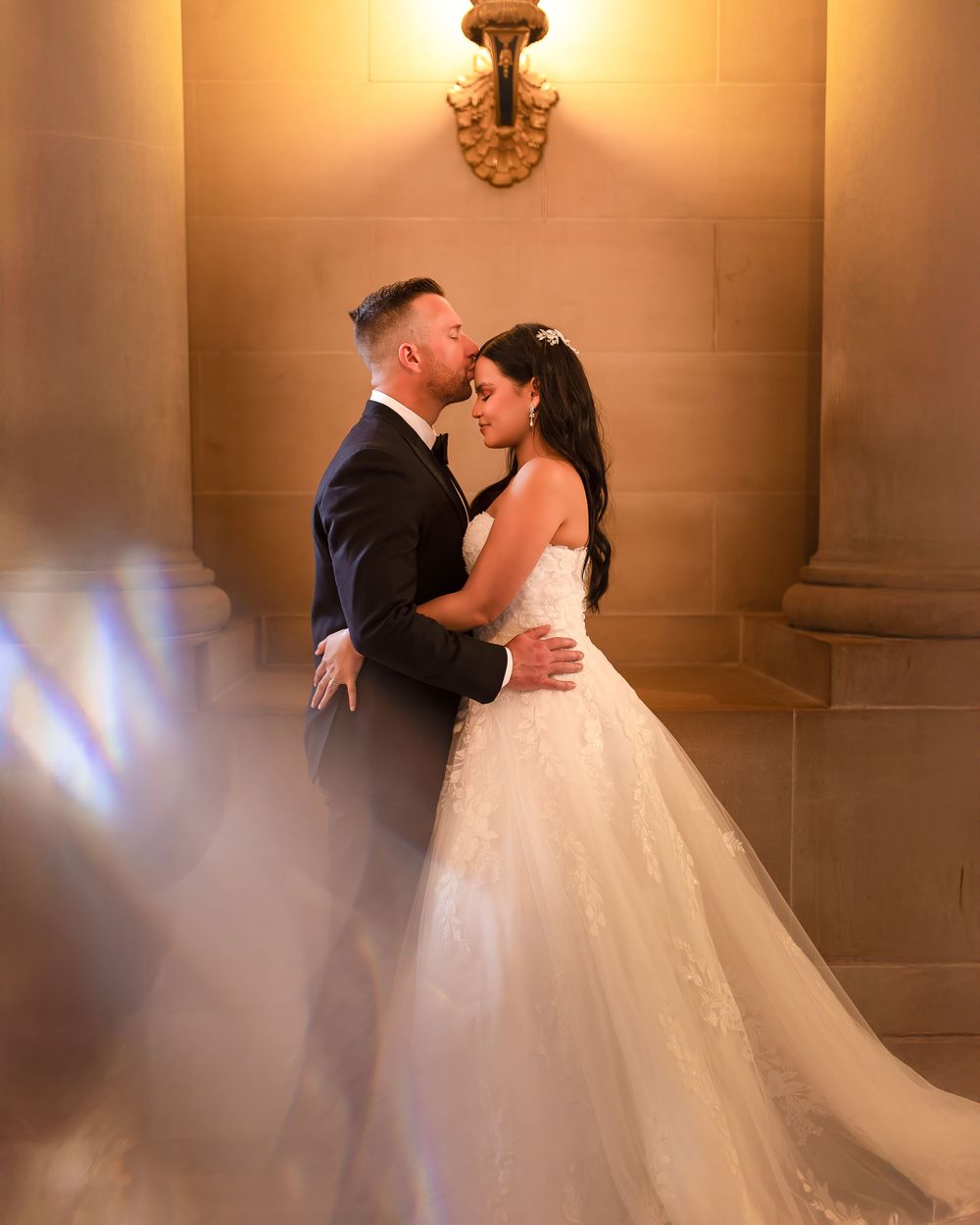 Bride and groom share a romantic embrace in an elegant venue, with warm golden lighting and ornate stone columns.