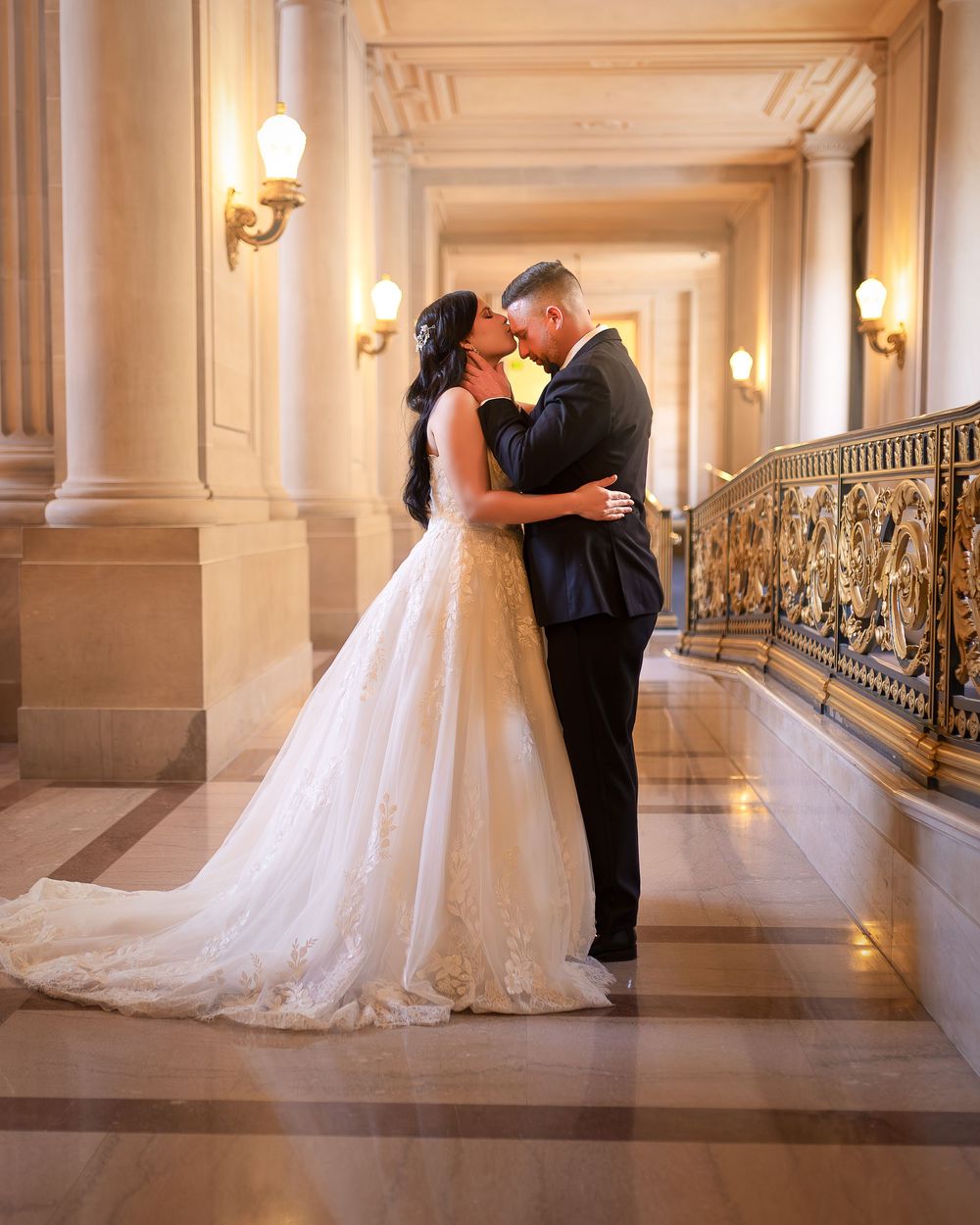 Bride and groom share a romantic kiss in an elegant marble hallway with ornate gold railings and warm glowing lights.