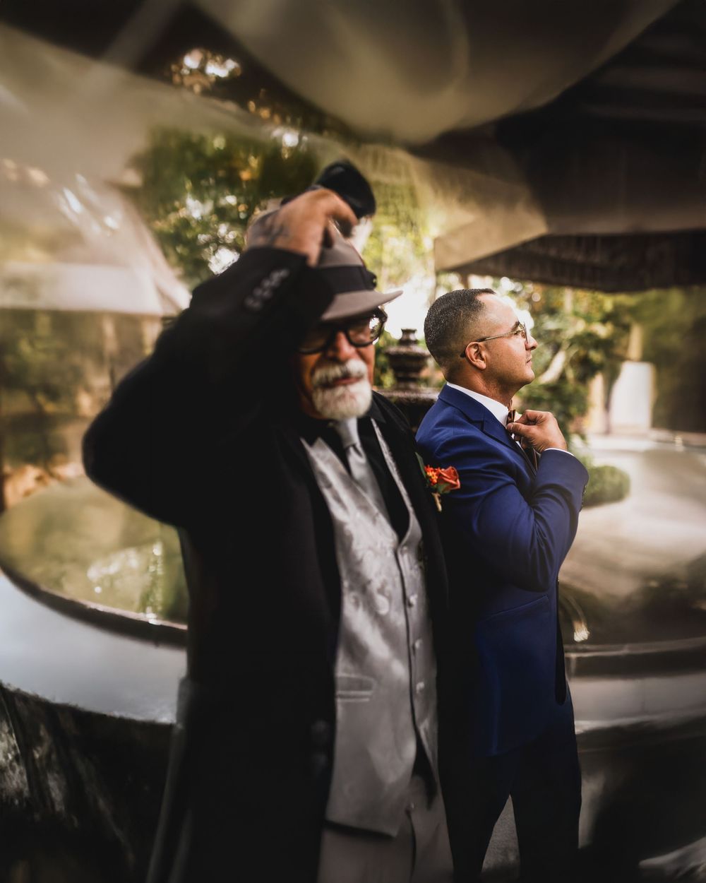 Two stylish men in formal attire, one in a gray suit with a fedora, another in a navy suit with a bow tie, pose outdoors.