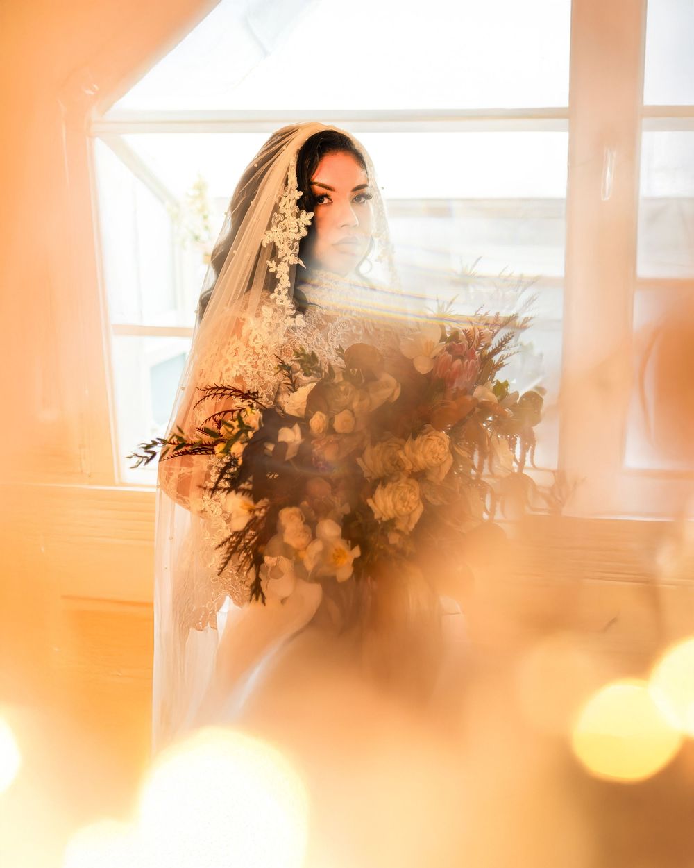 Bride in veil holding large floral bouquet by window with warm golden bokeh light.