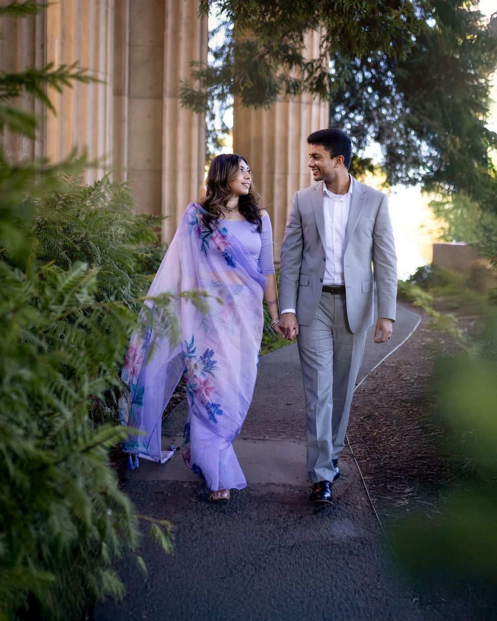 Couple walking together, woman in floral lavender saree and man in gray suit, strolling along a tree-lined path.