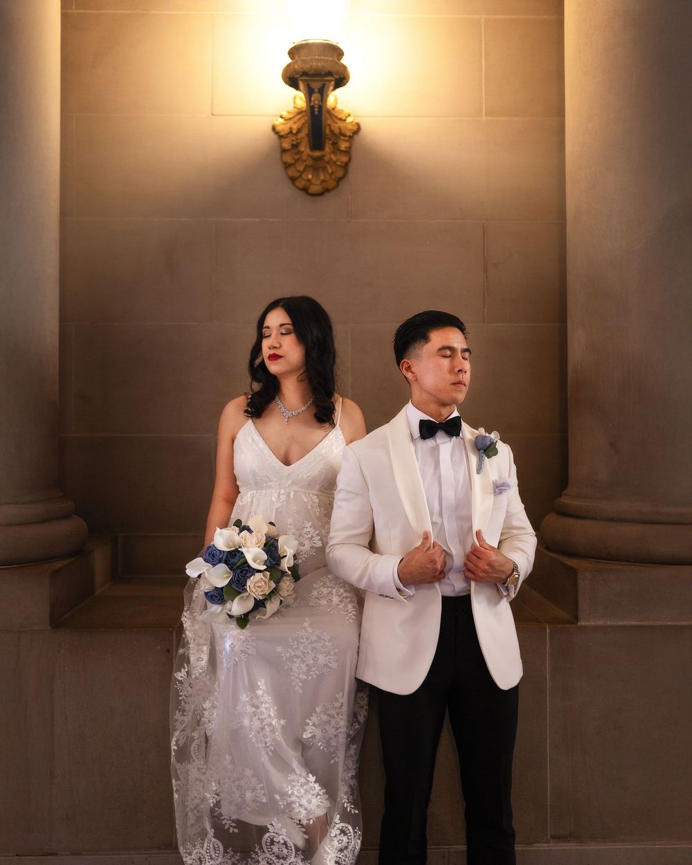 Elegant bride in lace gown holding bouquet stands with groom in white tuxedo jacket at a grand venue.