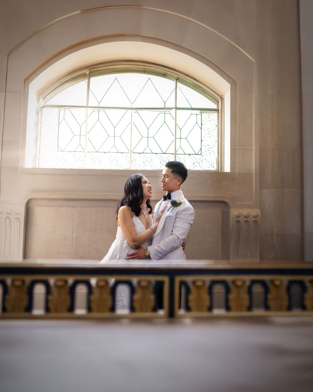 A bride and groom share a romantic moment in front of an arched stained glass window at an elegant wedding venue.