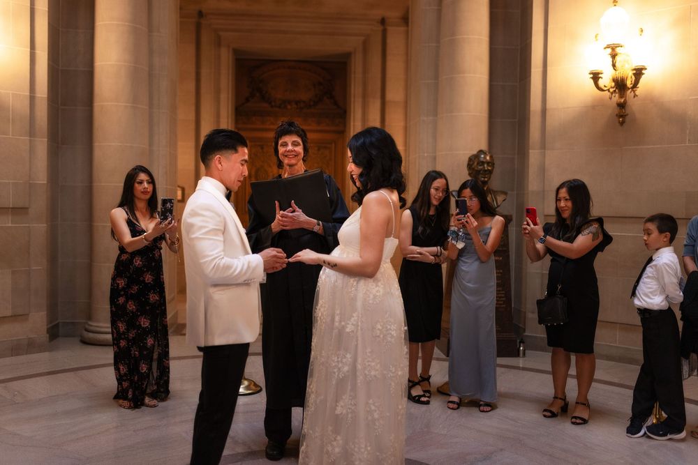 Couple exchanging vows at San Francisco City Hall wedding ceremony with guests watching and officiant in elegant marble rotunda.