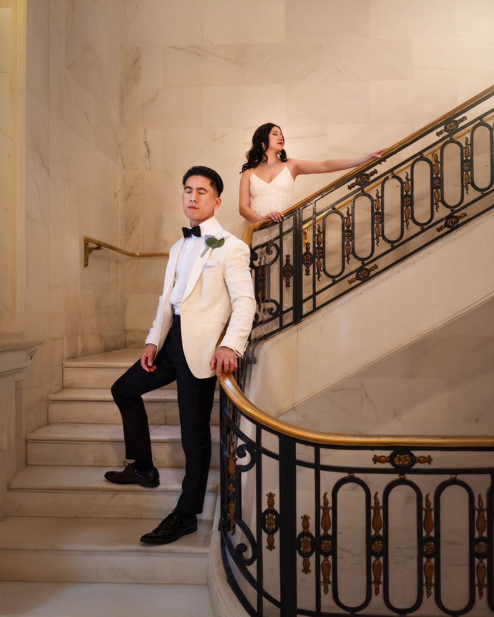Elegant wedding couple posing on ornate marble staircase, groom in white tuxedo and bride in white gown.