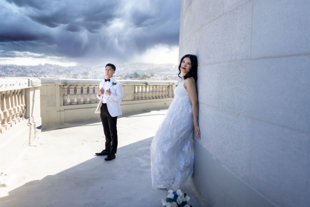 Bride in white gown leans against wall while groom in tuxedo stands on rooftop terrace under dramatic stormy sky.