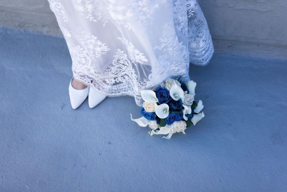 Bride in white lace wedding dress holding blue and white floral bouquet with white heels on blue surface.
