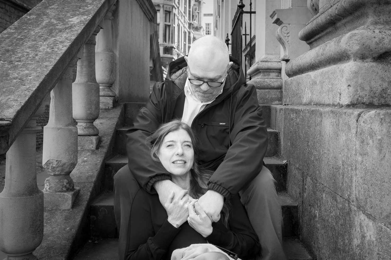 Black and white photo of a bald man and young girl sitting together on stone steps, sharing a warm embrace.