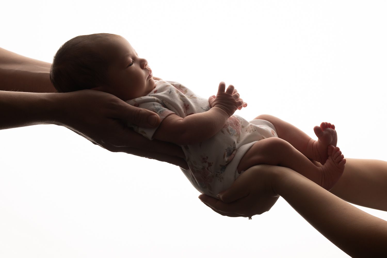 Silhouetted adult hands gently holding a sleeping newborn baby in soft white backlit light.
