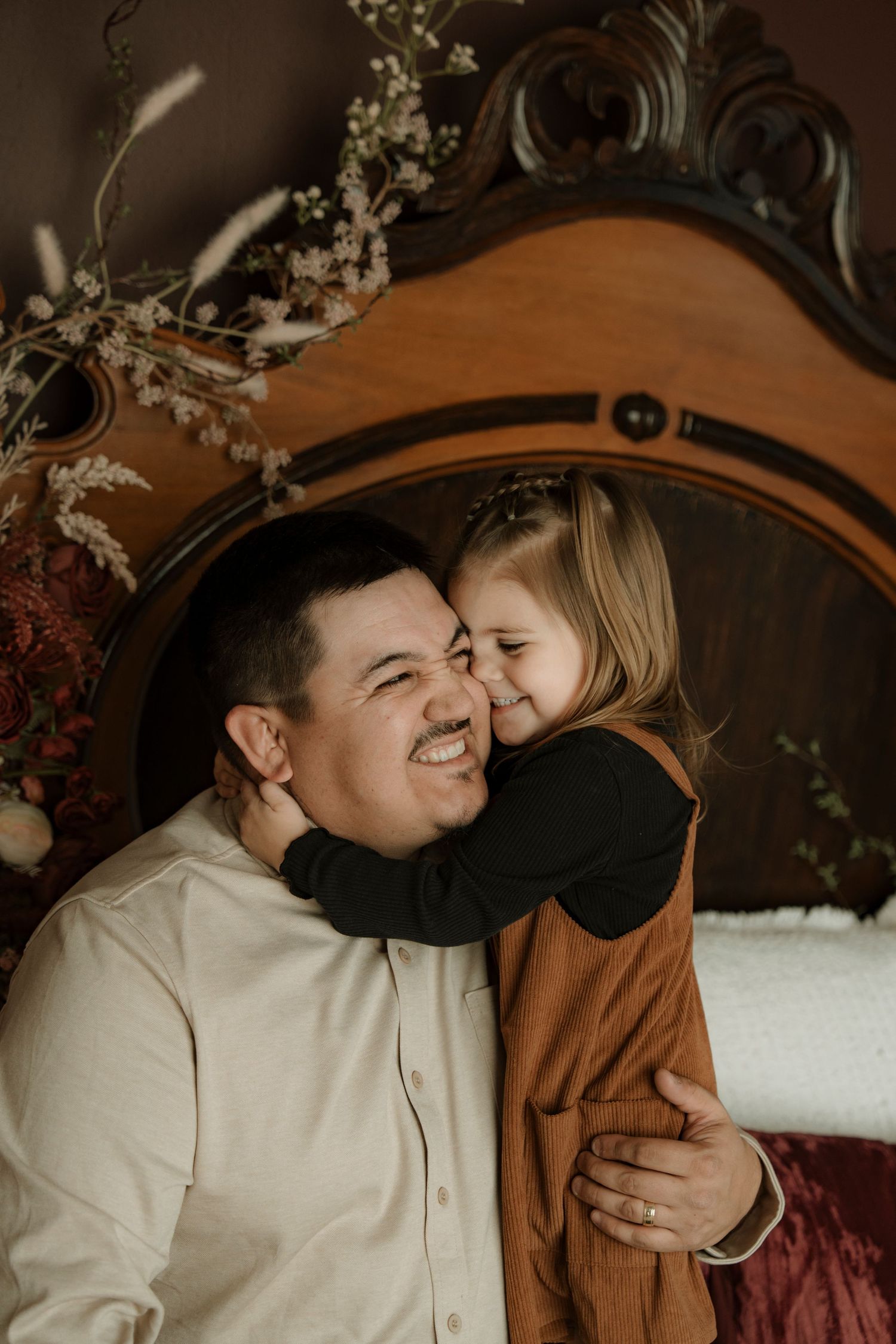 A laughing man hugs a young girl in a warm indoor portrait with vintage wooden headboard and floral decor.