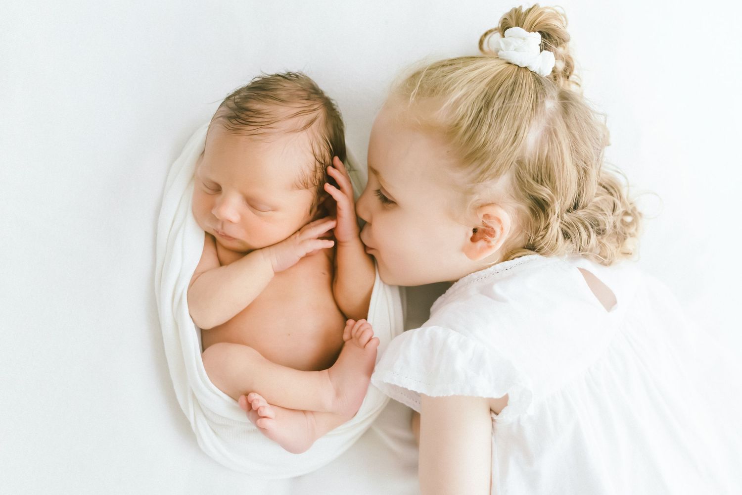 A toddler girl in a white dress gently kisses a sleeping newborn baby wrapped in a white swaddle.