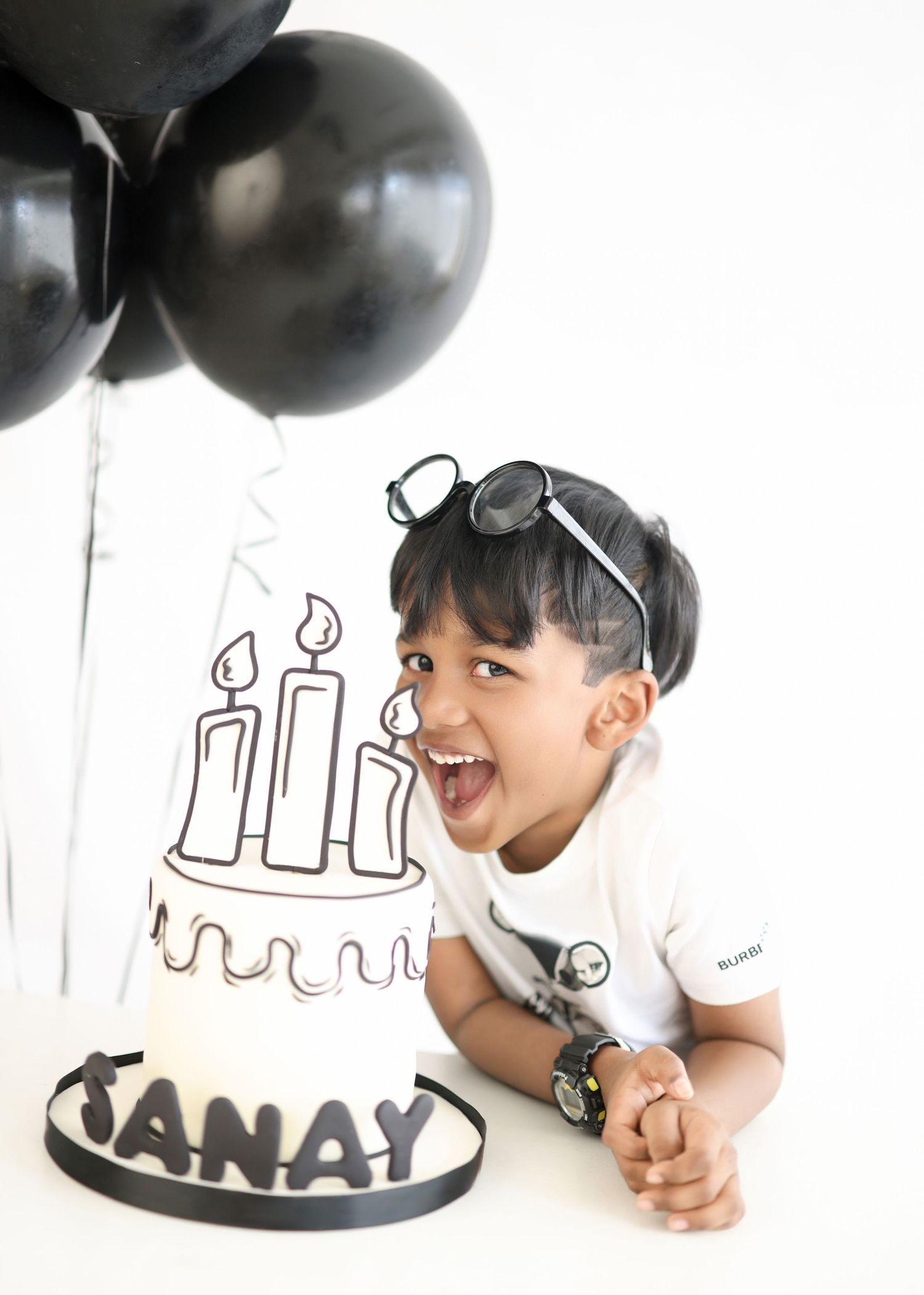 Joyful toddler named Janay laughing beside a birthday cake with black balloons and sunglasses on head.