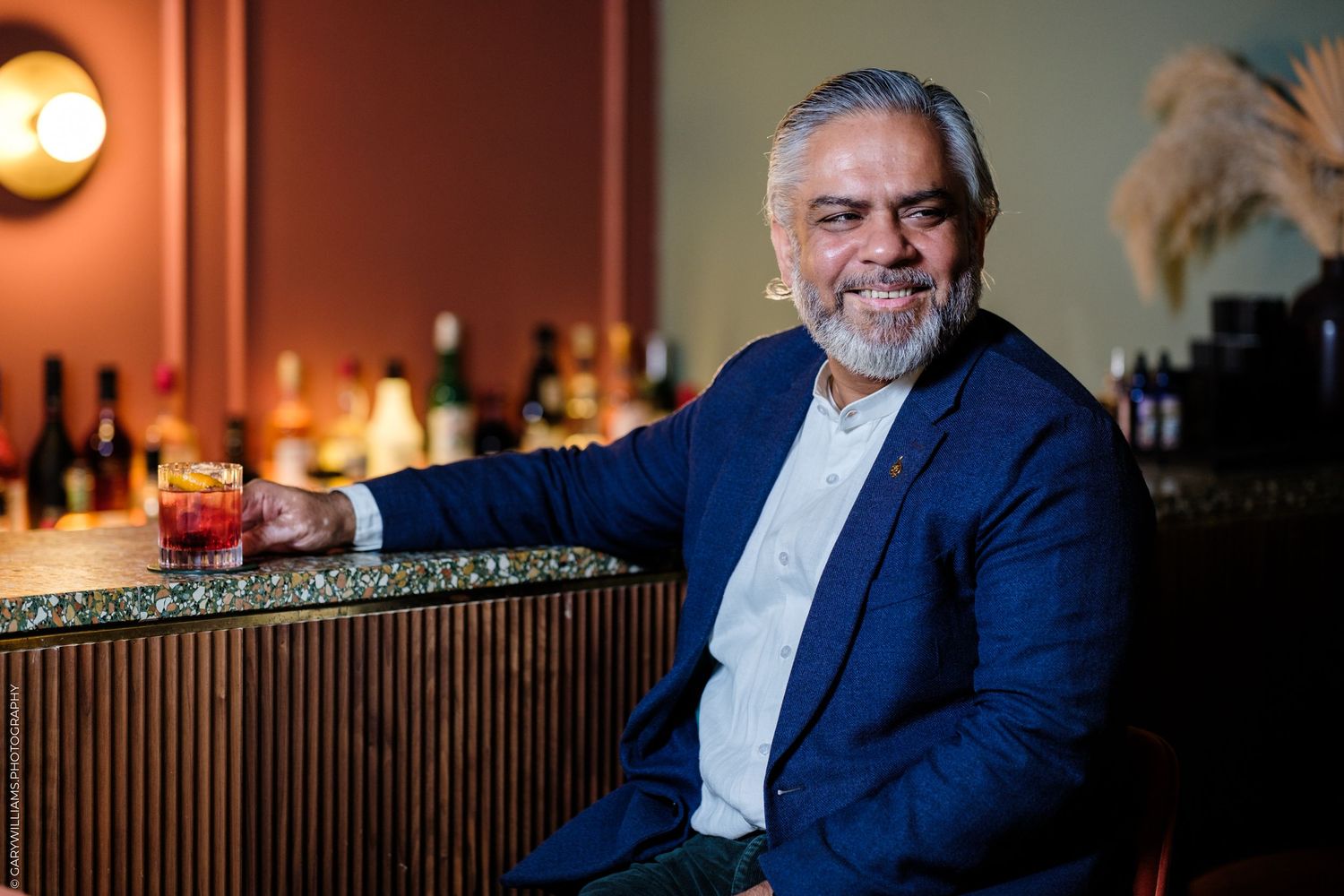 Smiling man in blue blazer holding cocktail glass at upscale bar with warm lighting and bottle display.