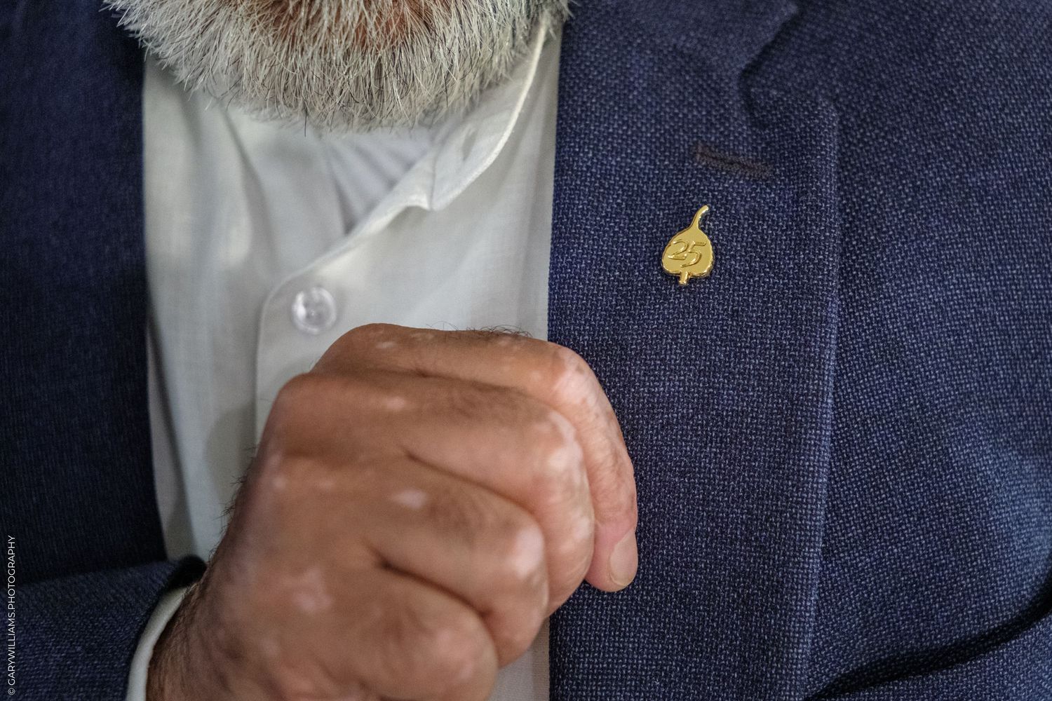 Close-up of an elderly bearded man in a navy suit with a gold leaf lapel pin, adjusting his jacket button.