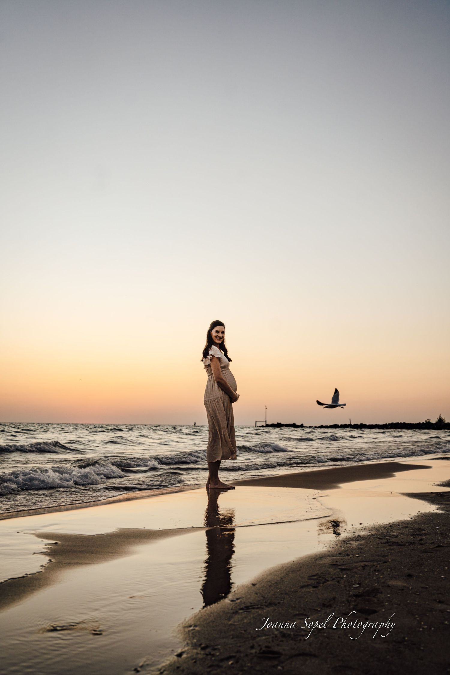 Pregnant woman in beige dress standing on beach at sunset with seagull flying over ocean waves.