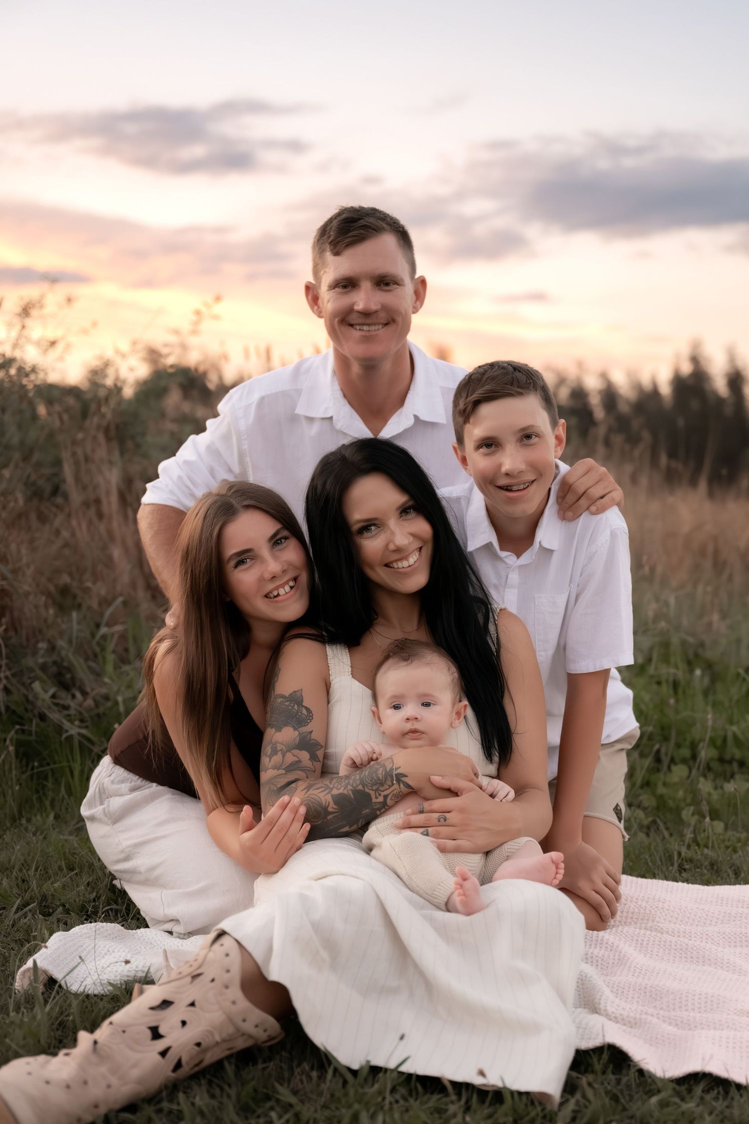 Happy family of five posing together on grass at sunset with beautiful pink and purple sky.
