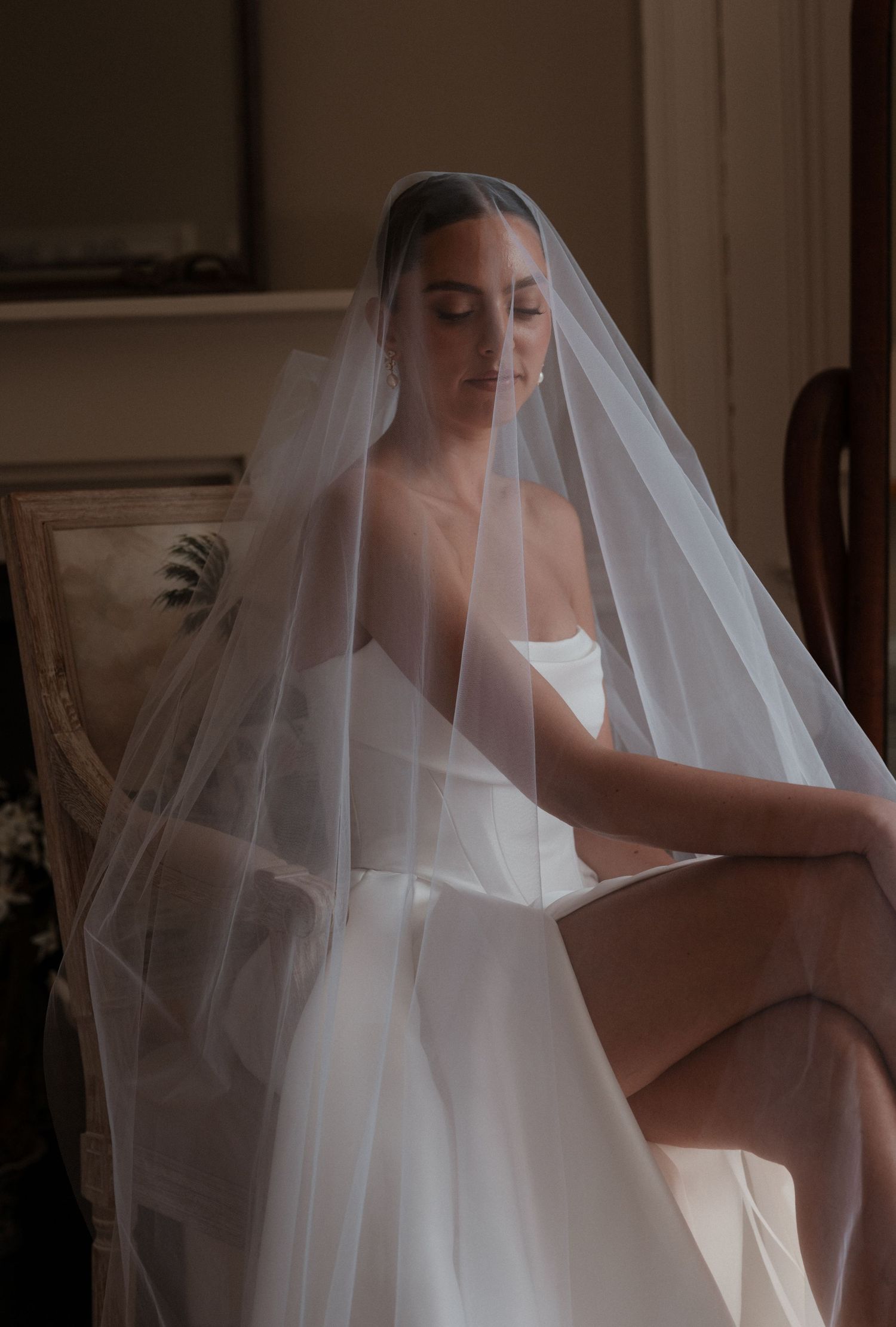 Bride in strapless white wedding dress seated gracefully, draped in flowing tulle veil, soft romantic lighting.