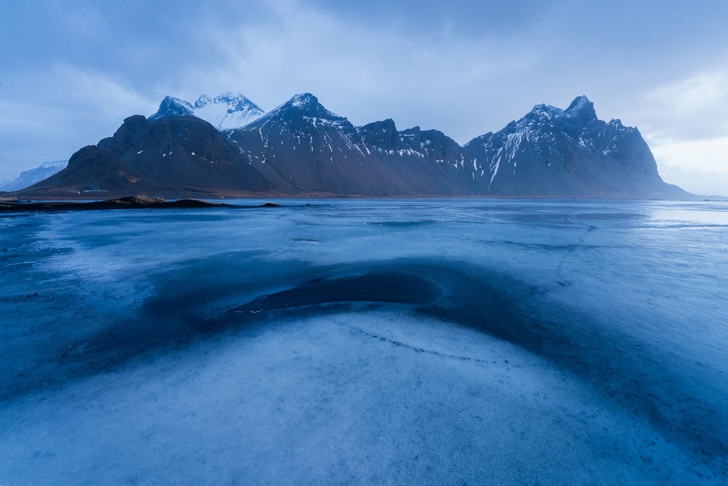 Frozen blue lagoon reflecting snow-capped Vestrahorn mountain range under moody winter twilight in Iceland.
