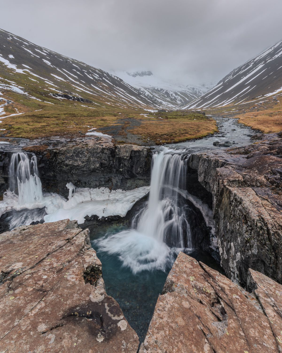 Twin waterfalls cascading into turquoise pool within a snow-dusted Icelandic mountain valley.