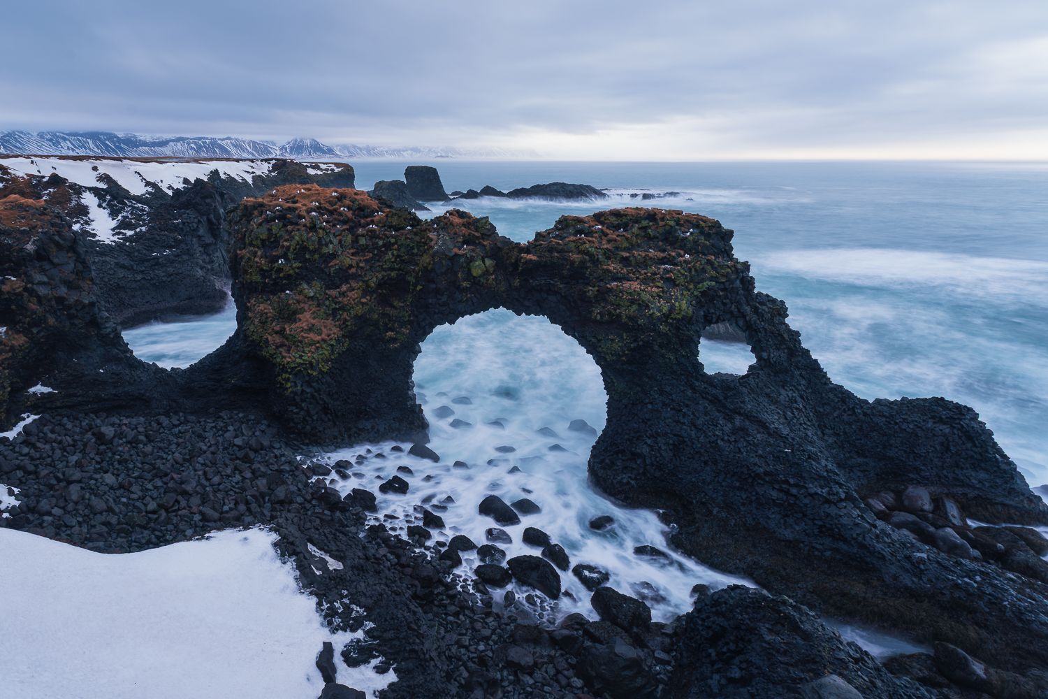 Ancient black lava rock arch formation over crashing winter waves on Iceland's snowy coastline.