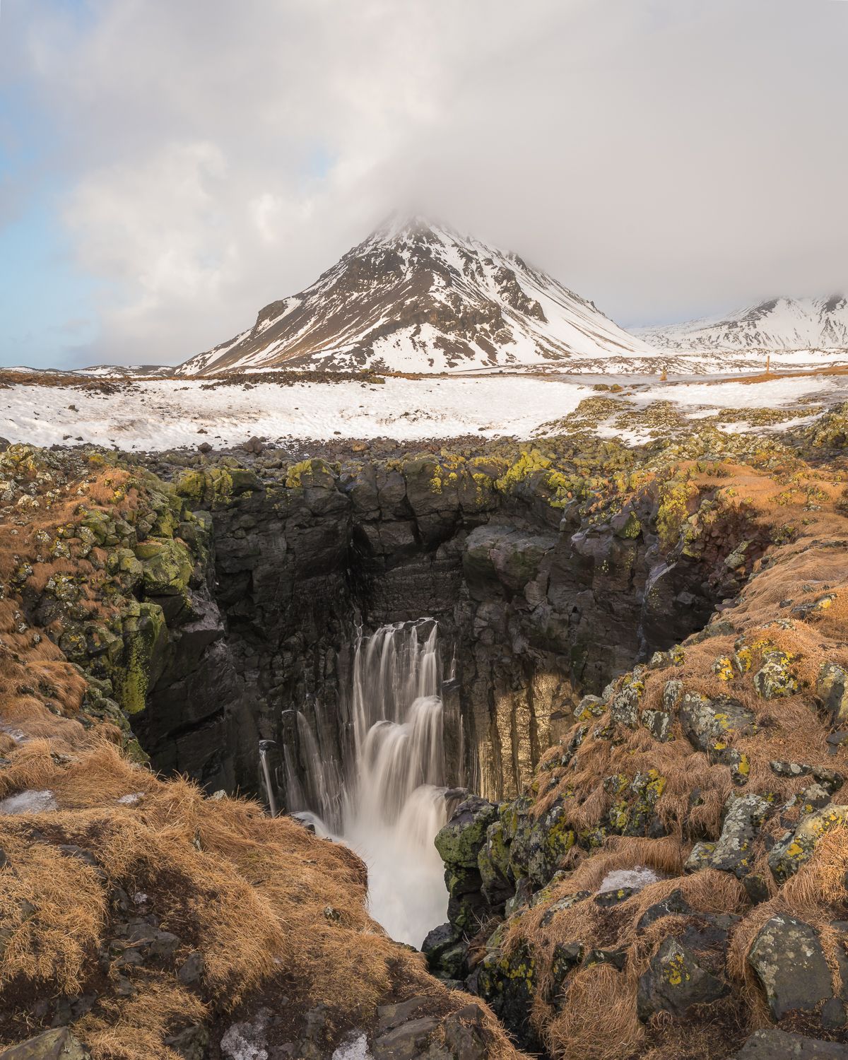 Silky waterfall plunging into rocky gorge with snow-capped volcanic mountain under cloudy sky in Iceland.