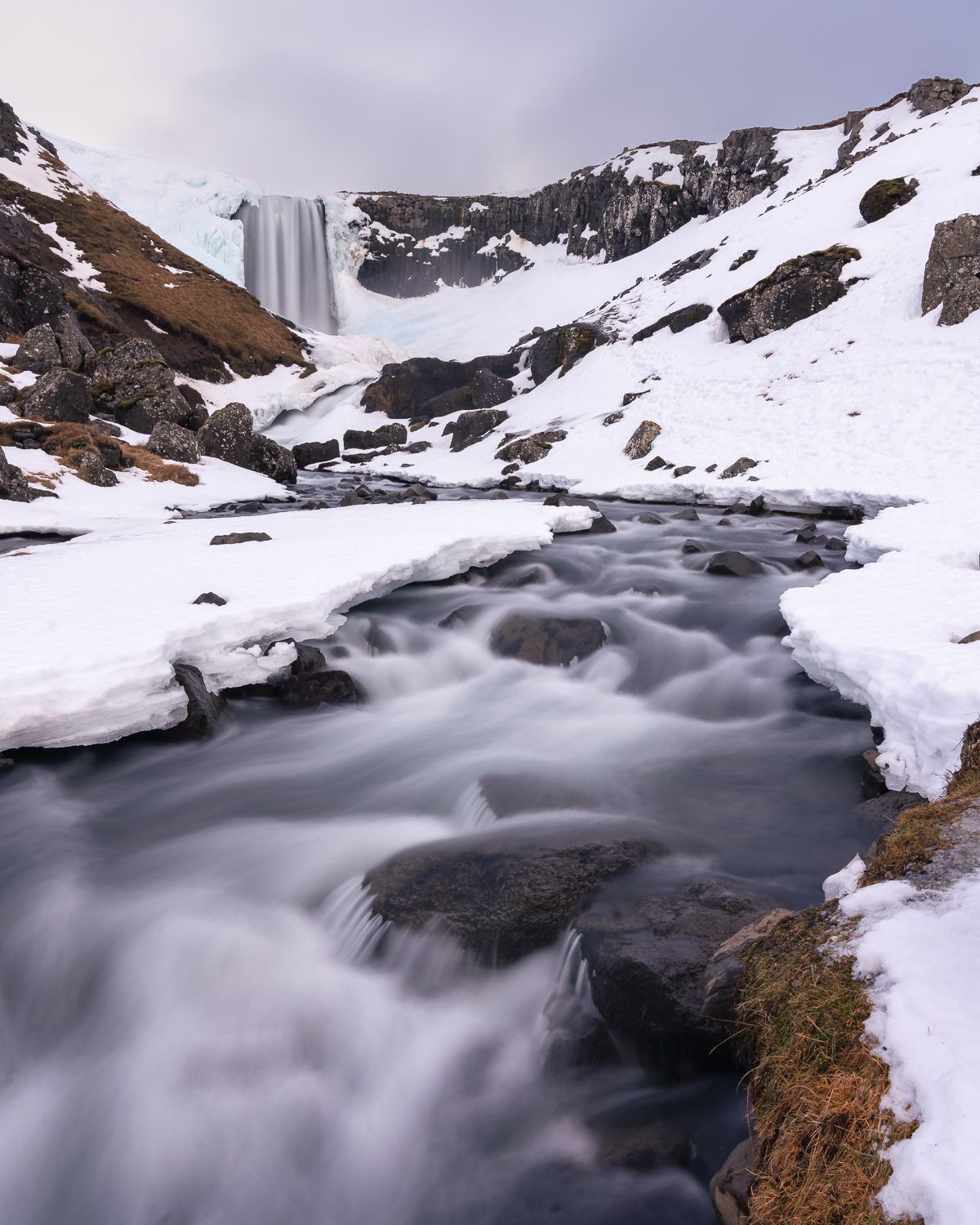Frozen waterfall and silky stream flowing through snow-covered rocky valley in Iceland's winter wilderness.