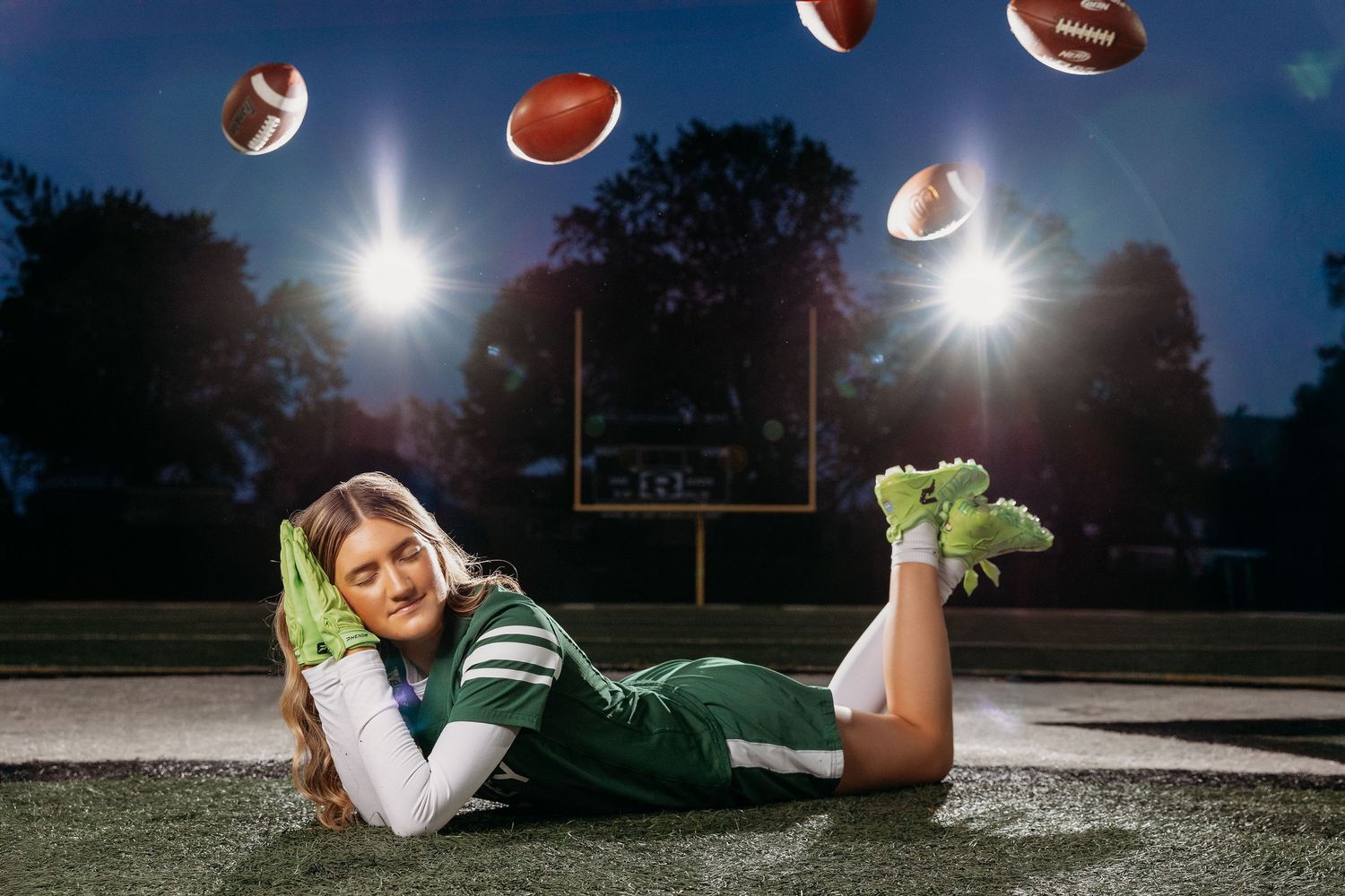 Cheerleader in green uniform poses on football field at night with footballs flying through the air.