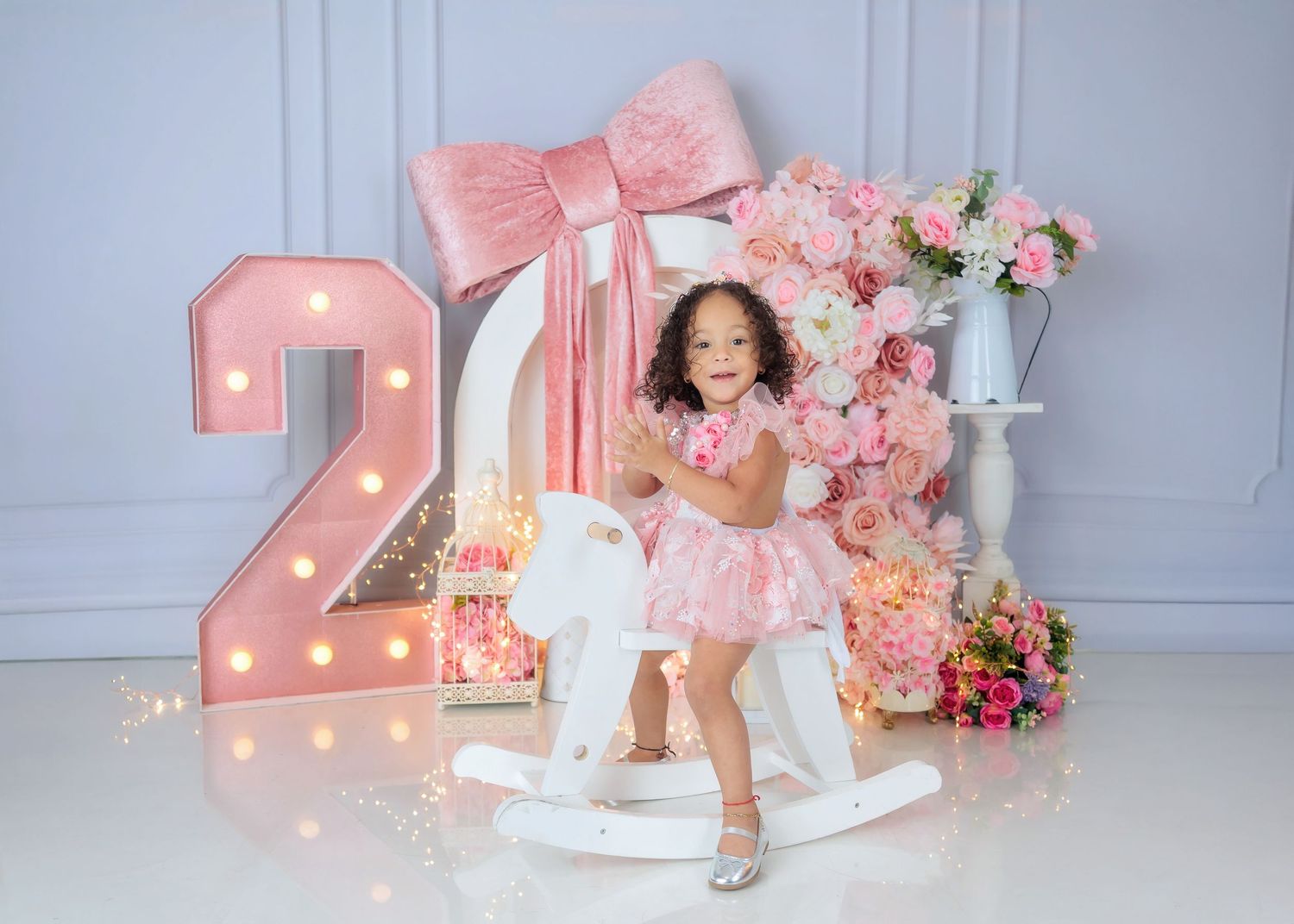 A toddler girl in a pink dress sits on a white rocking horse beside a marquee number 2 and floral decorations.