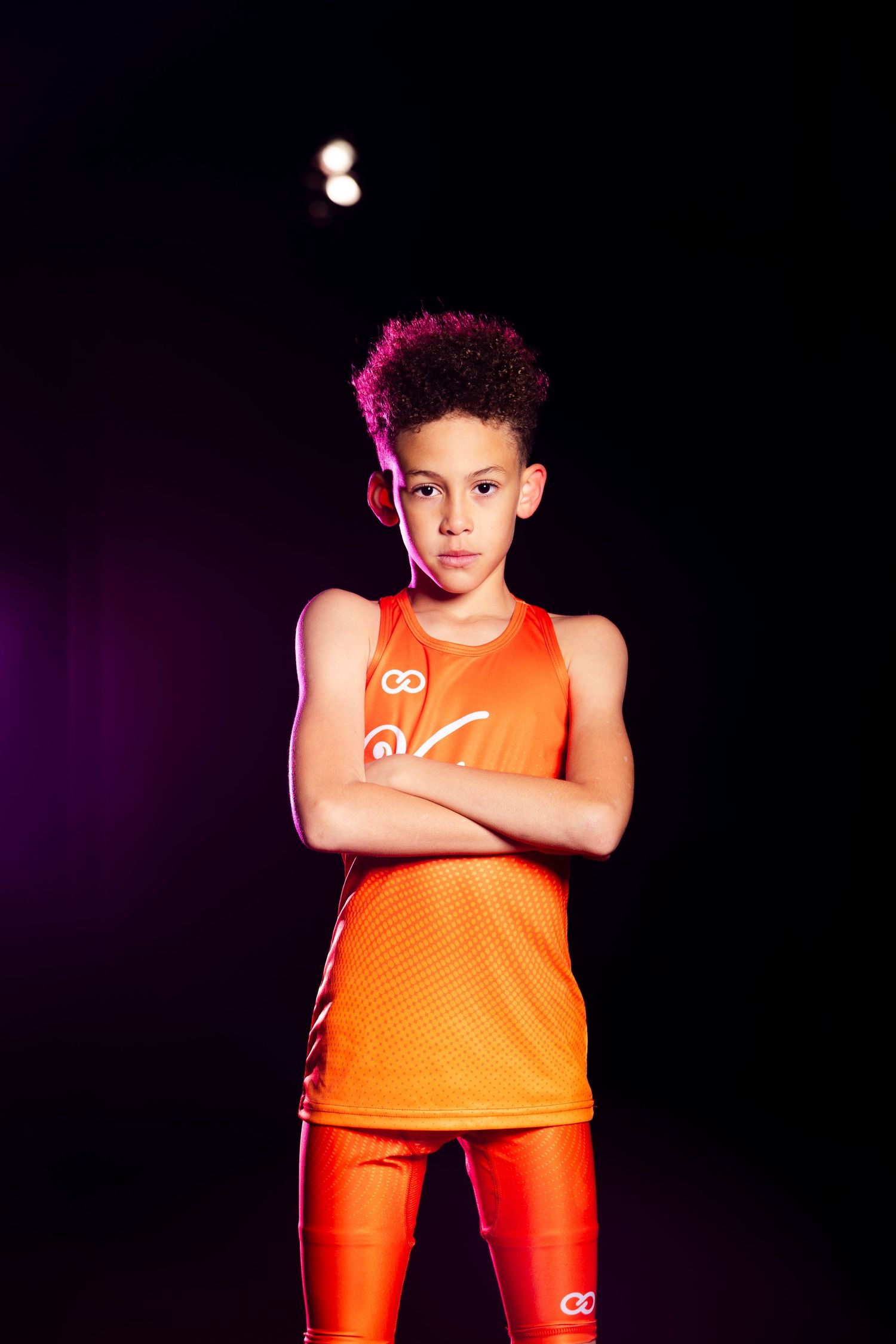 Young athlete in orange sports uniform stands confidently with arms crossed against dark background.