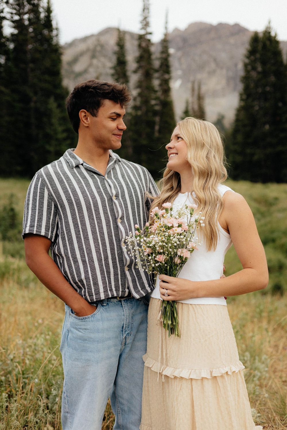Couple sharing a romantic moment in a mountain meadow, woman holding wildflowers bouquet.