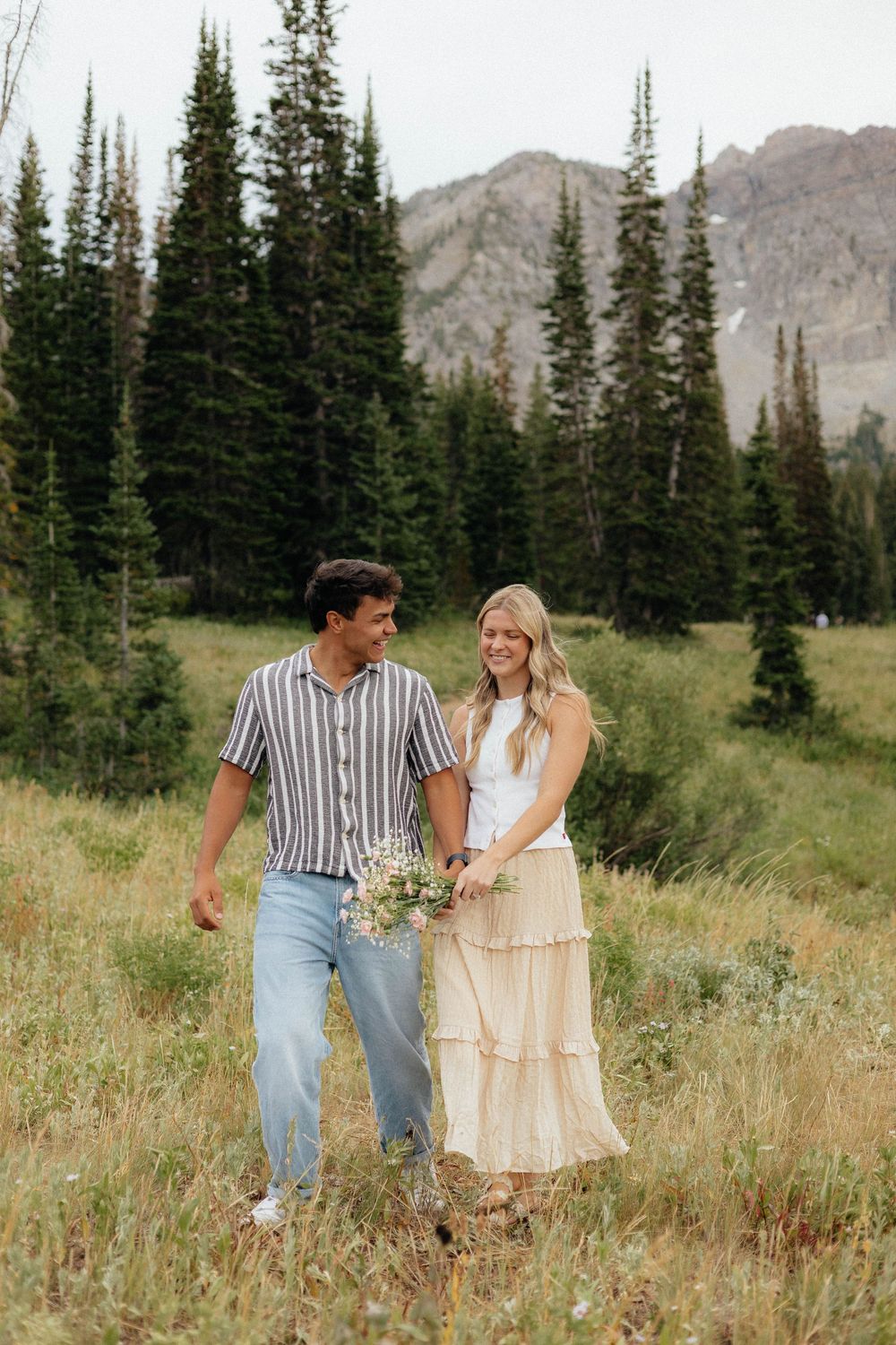 Happy couple walking hand-in-hand through alpine meadow with evergreen trees and mountains.