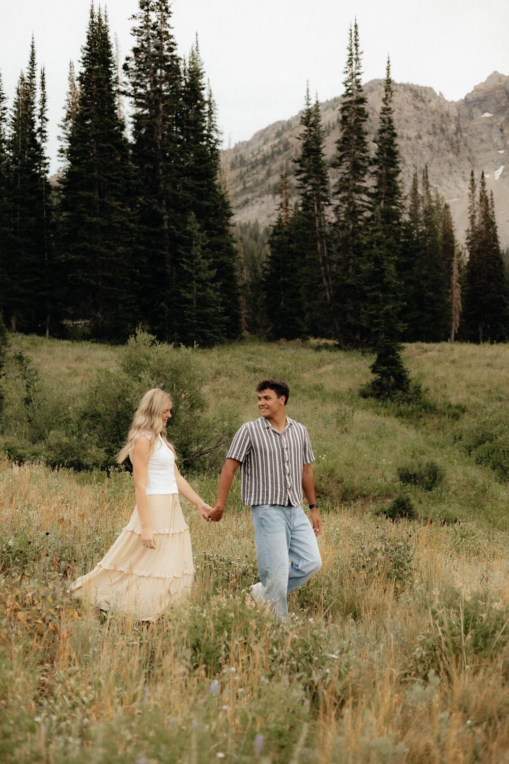 Couple holding hands walking through a wildflower meadow with pine trees and mountains in the background.