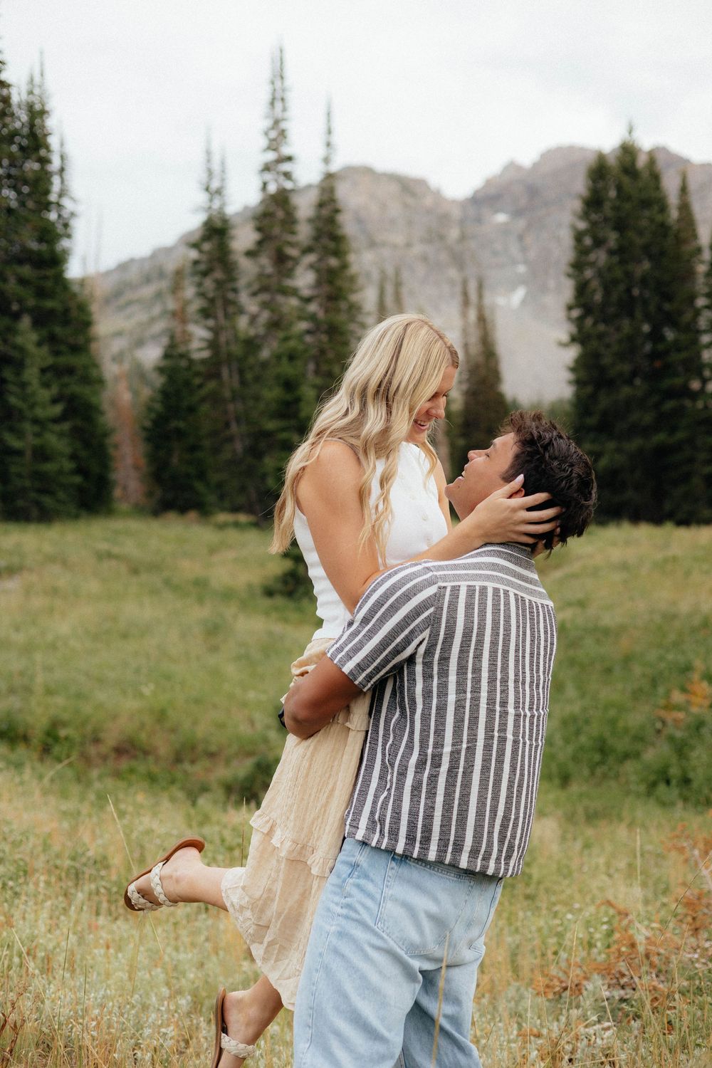 Man lifting woman in alpine meadow with pine trees and mountains, romantic couple engagement photo outdoors.