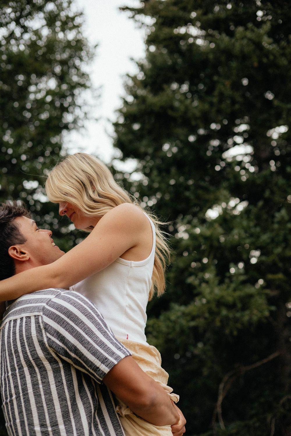 Man lifting smiling blonde woman against evergreen trees in a tender romantic outdoor couples moment.