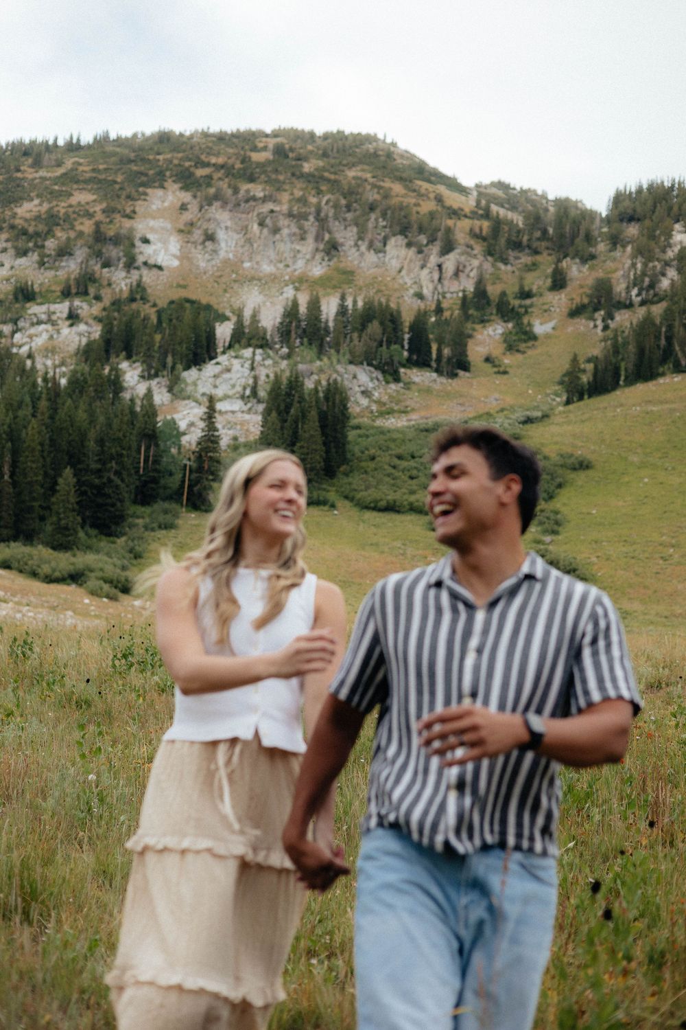 Happy couple holding hands laughing in mountain meadow with rocky peaks and pine trees behind them.