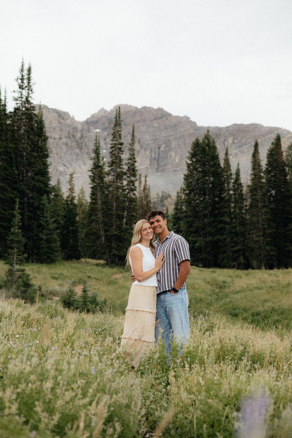 Couple embracing in a wildflower meadow with pine trees and rocky mountains in the background.