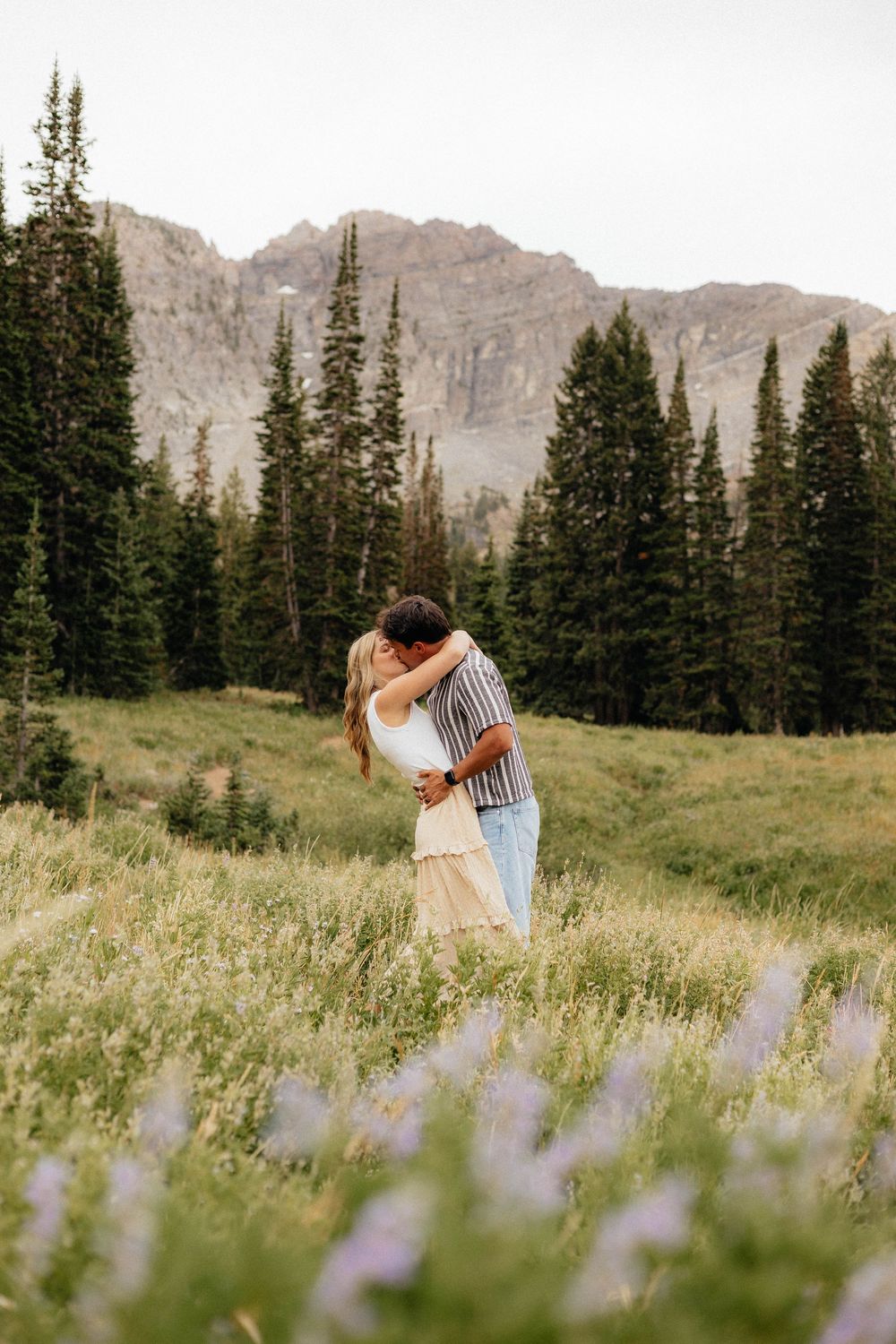 Couple kissing in a wildflower meadow with pine trees and mountain backdrop.