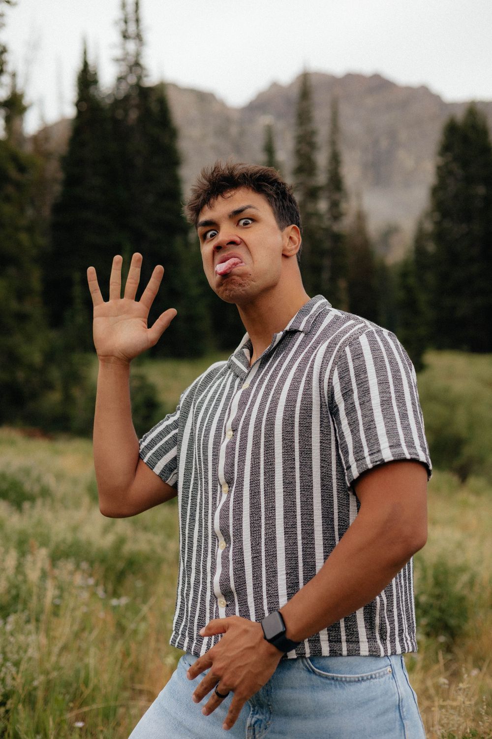 Young man in striped shirt making funny face while raising hand outdoors with mountain and pine trees background.