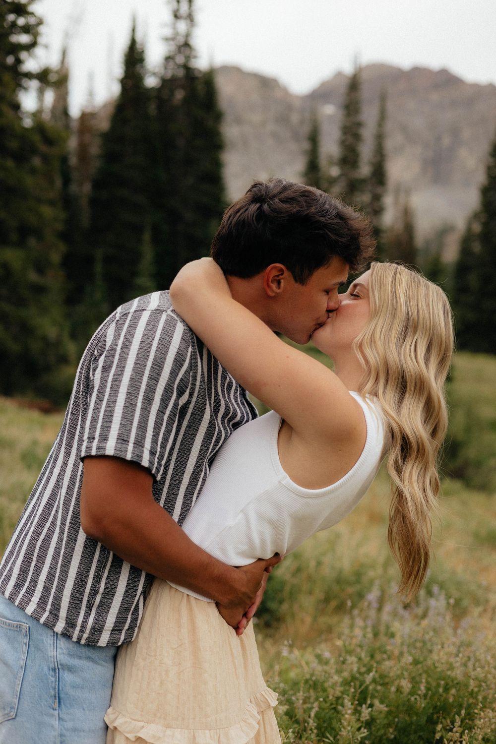 Couple sharing a romantic kiss in a lush alpine meadow with evergreen trees and mountains behind them.