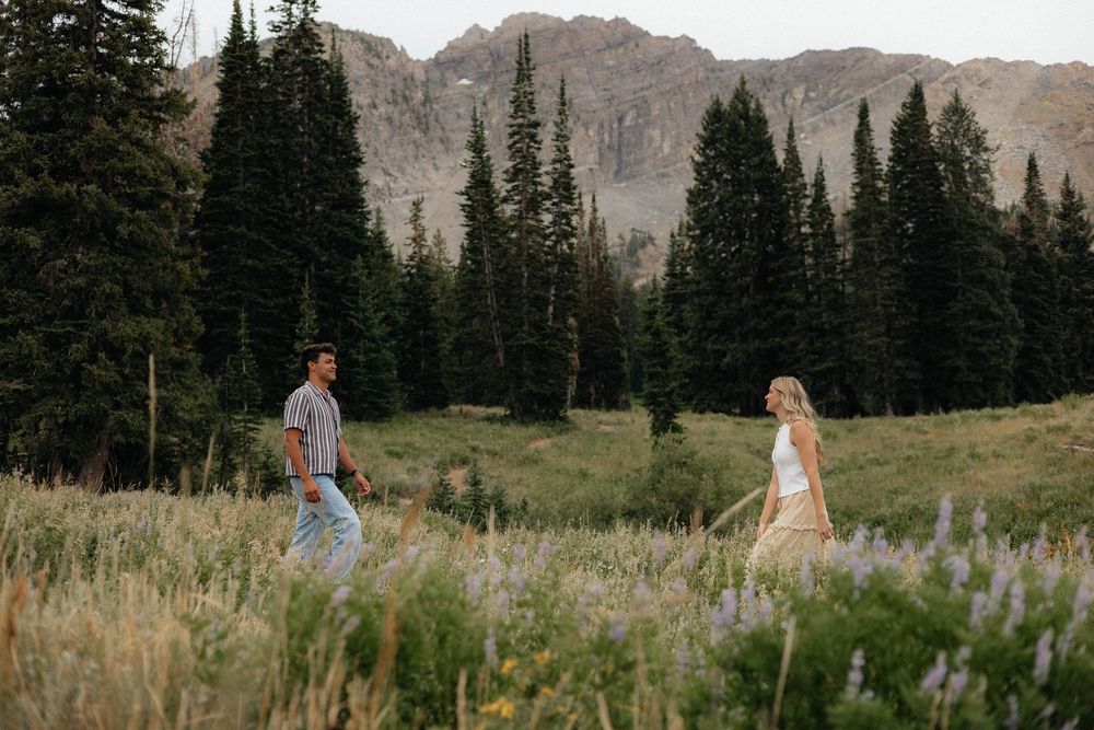 Couple walking through alpine wildflower meadow with pine trees and mountains.