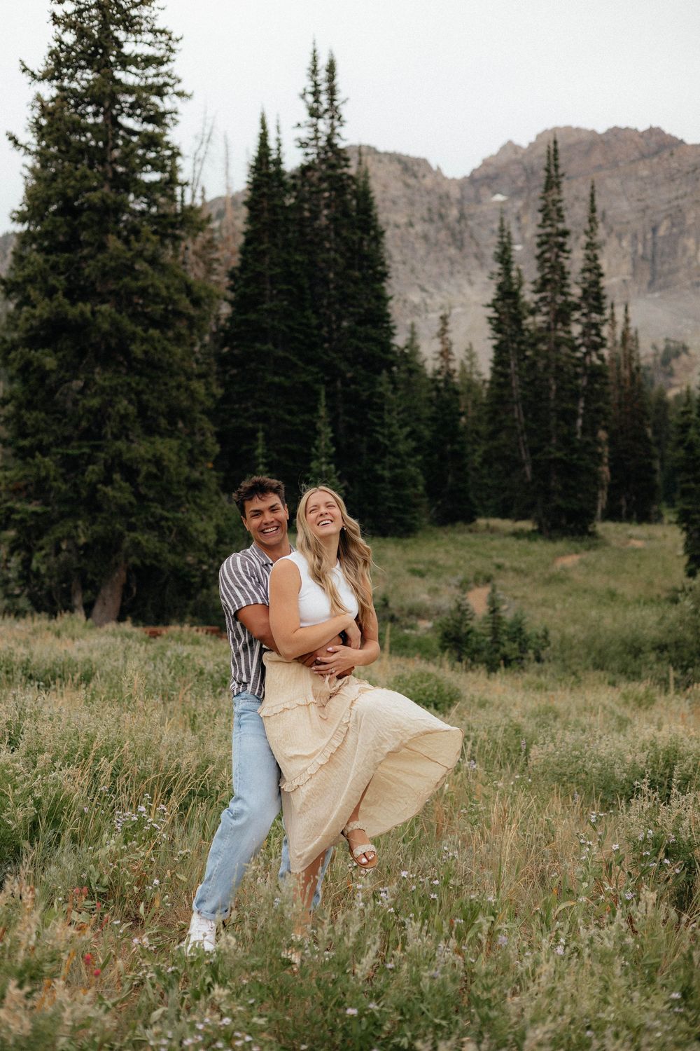 Laughing couple embracing in a mountain meadow with evergreen trees behind them.